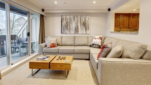 A modern living room with a sectional sofa, a wooden coffee table, and a large window offering a view of the outdoors. - Heavenly 2BR/2BA. Private Hot-tub & Mountain View’s (Whistler)
