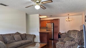 Sala de estar con sofá, sillón y mesa de centro. Se observa una zona de cocina con armarios y refrigerador. Hay un ventilador de techo y una lámpara de araña.