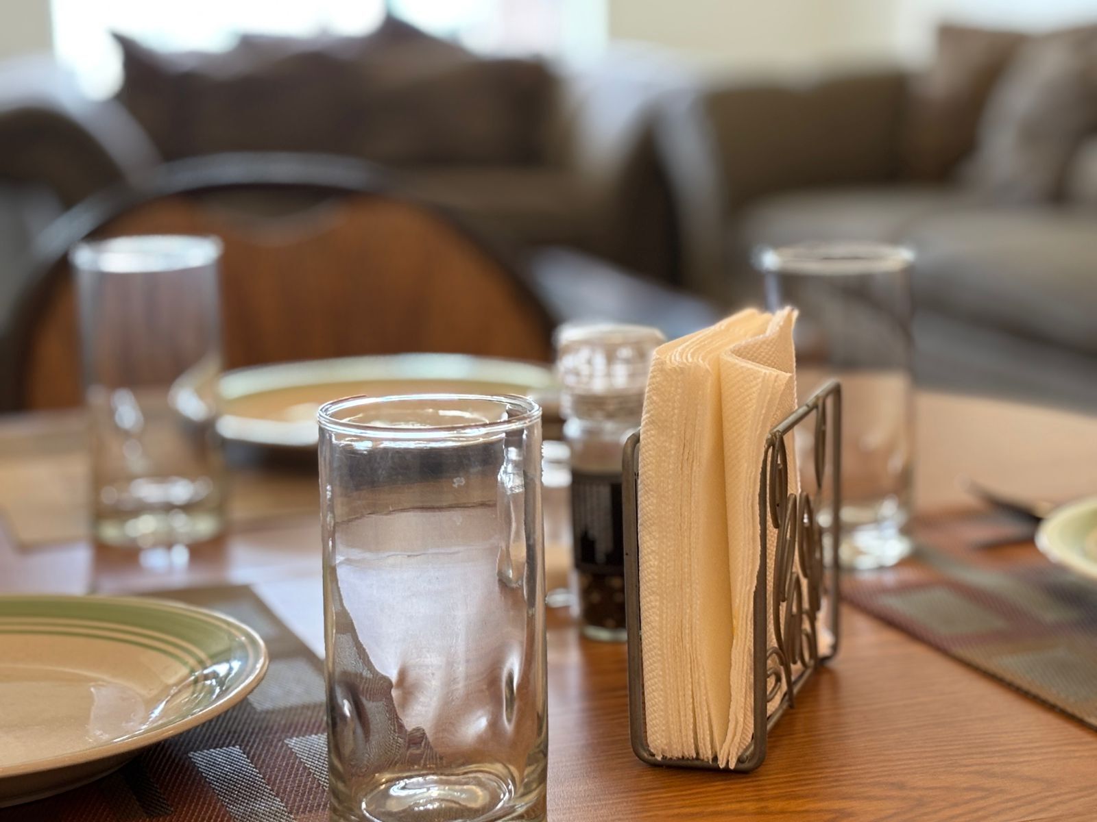 A dining table with clear glassware, a napkin holder, and plates, set in a room with a sofa and cushions in the background.