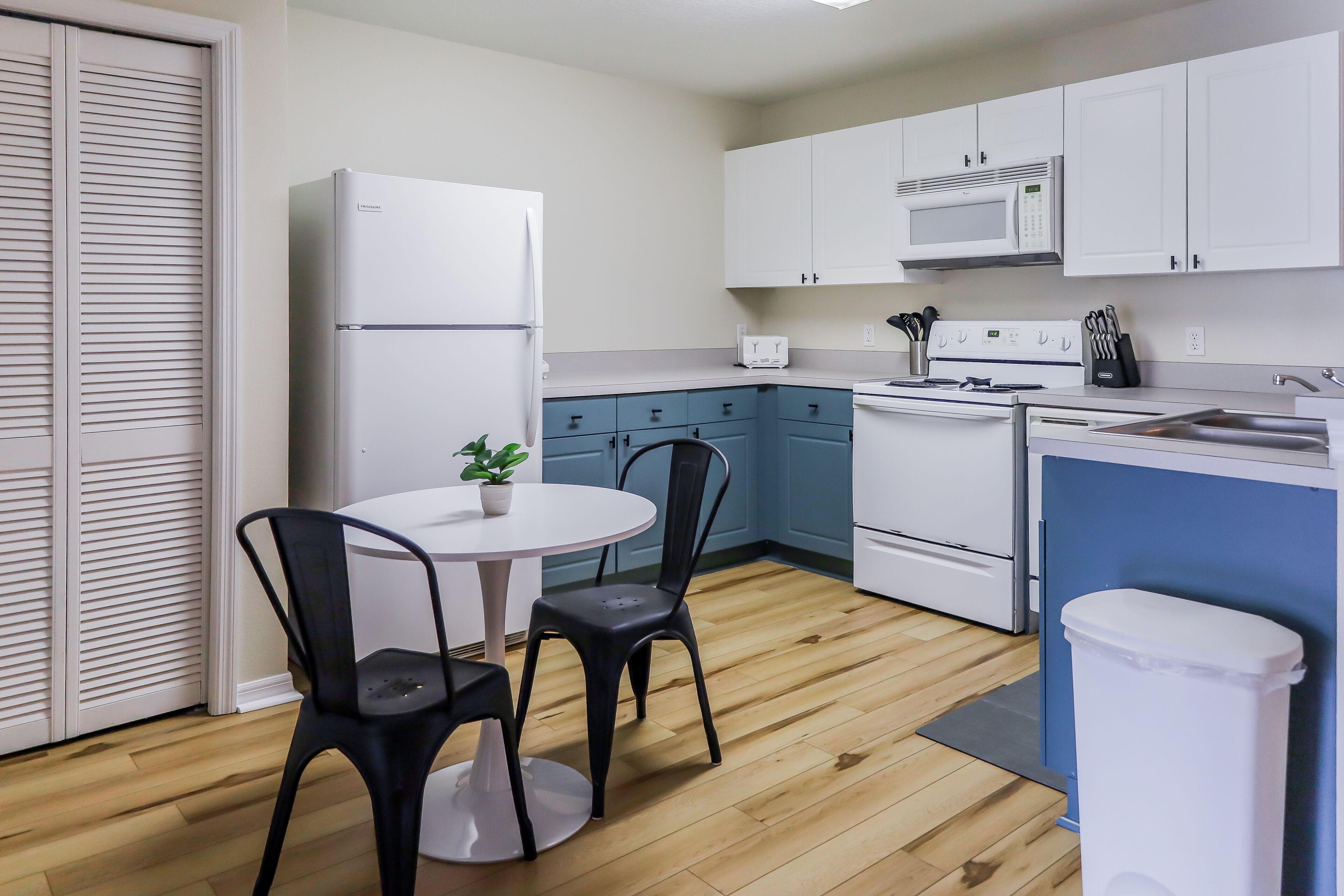 Une cuisine compacte avec un réfrigérateur blanc, un four à micro-ondes, une cuisinière et un évier. On y trouve une petite table à manger avec deux chaises et une plante en pot.