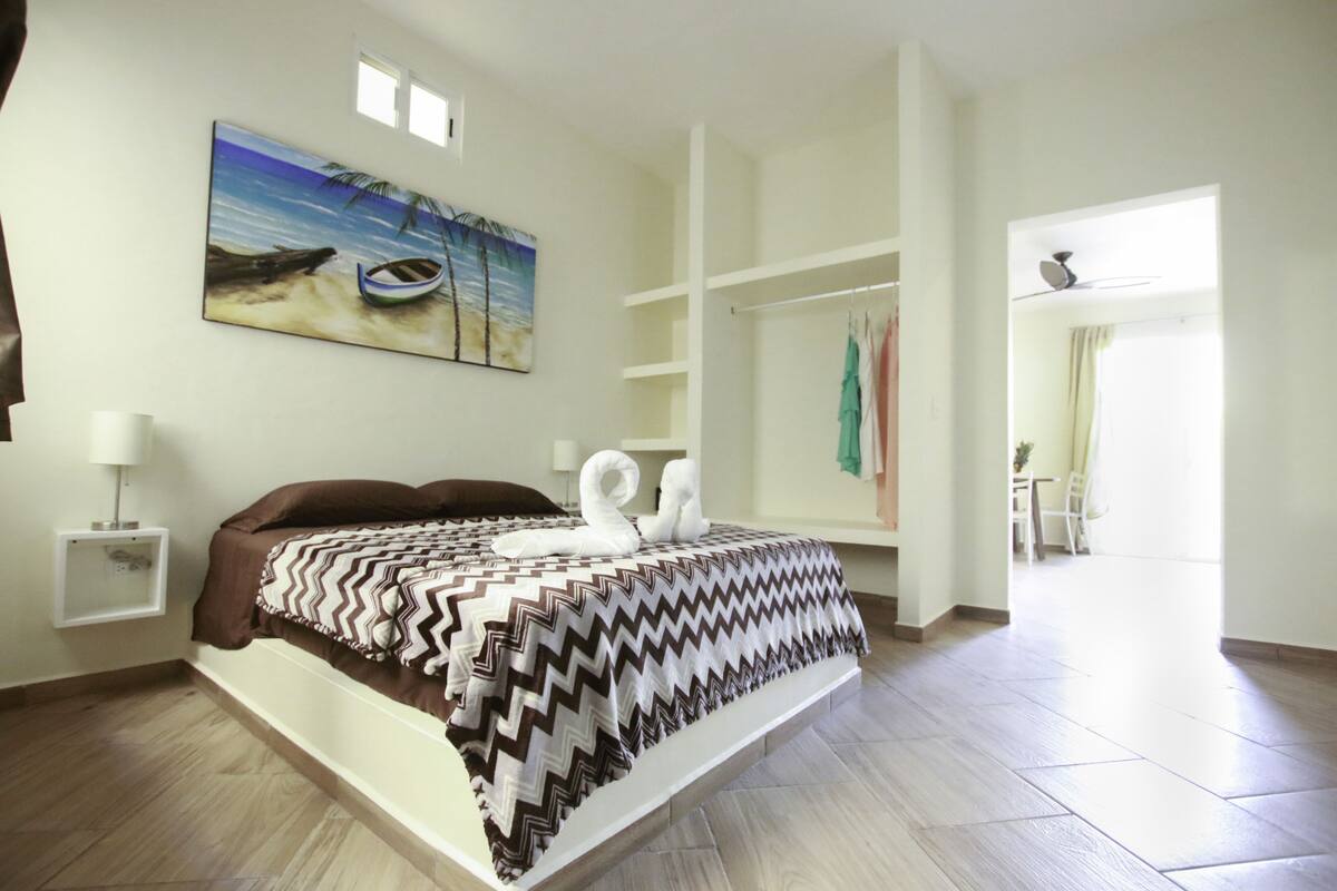 A bedroom with a bed, a painting of a beach scene, a wardrobe, and a view to a dining area.