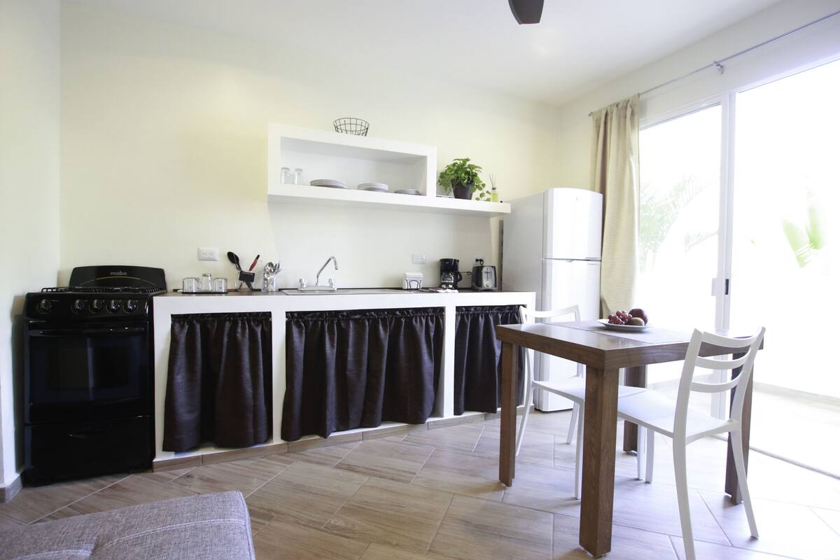 A compact kitchen with a stove, sink, and white cabinets. There is a dining table with chairs and a window with curtains.