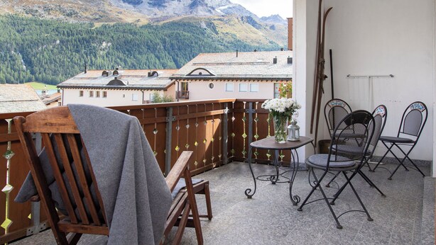 A balcony with a view of mountains, wooden chairs, a table with a vase of flowers, and metal chairs.