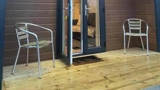 Une structure accueillante, partiellement ouverte, avec une façade en bois et une porte en verre donnant sur l’intérieur. Deux chaises en métal sont placées à l’extérieur.