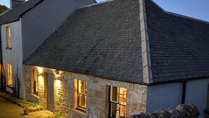 Une maison en pierre avec un toit en pente, des fenêtres illuminées et un mur de pierre au premier plan.