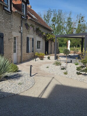 A stone building with a tiled roof, a gravel courtyard, and a pergola structure.