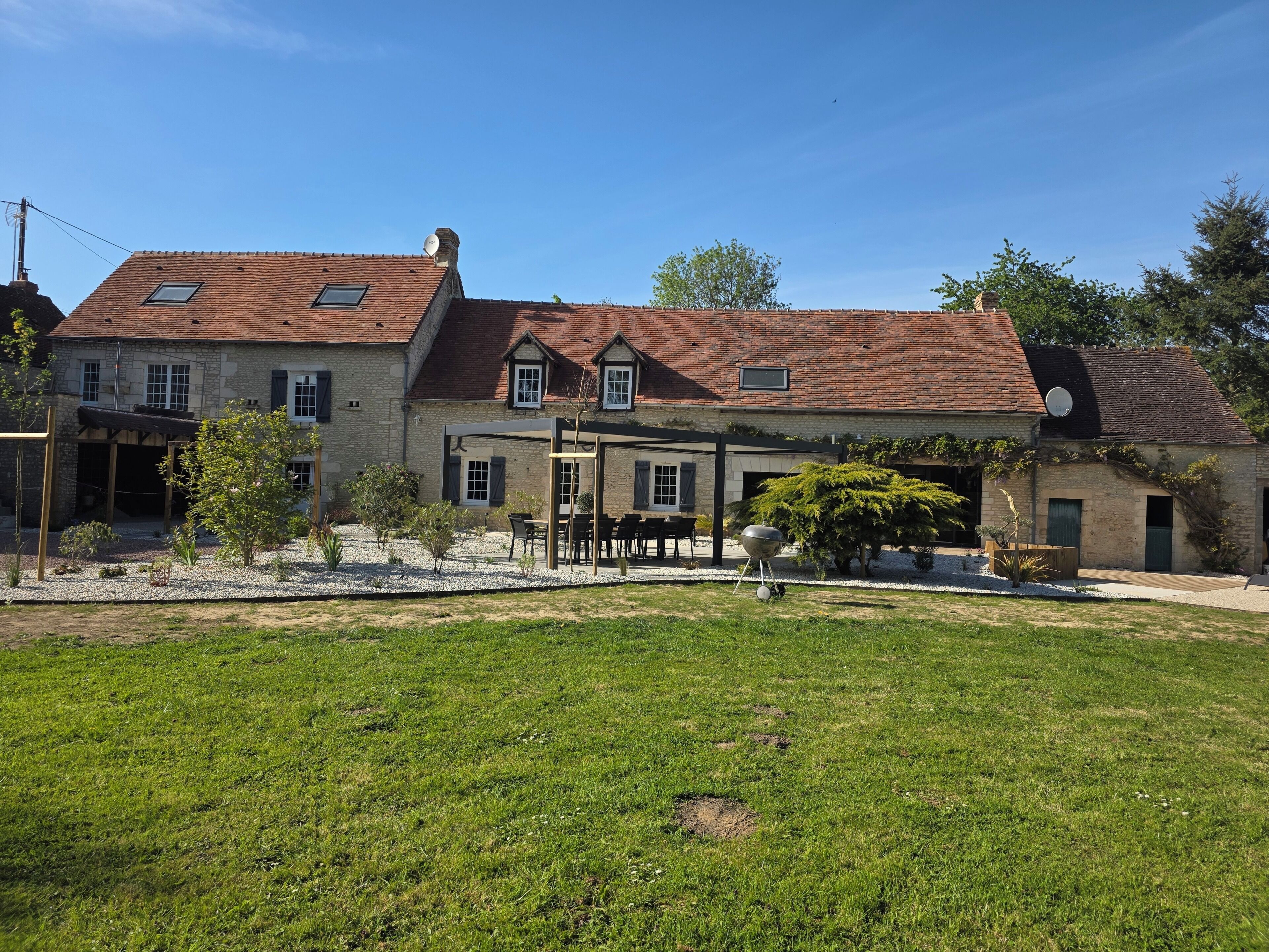 En bygning i sten med rødt tegltag, en overdækket terrasse med udendørs møbler, og en velplejet græsplæne.