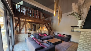 A spacious living area with a large sofa set, a coffee table, and a fireplace. There is a staircase leading to an upper level, and a dining area is visible in the background.