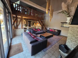 A spacious living area with a large sofa set, a coffee table, and a fireplace. There is a staircase leading to an upper level, and a dining area is visible in the background.