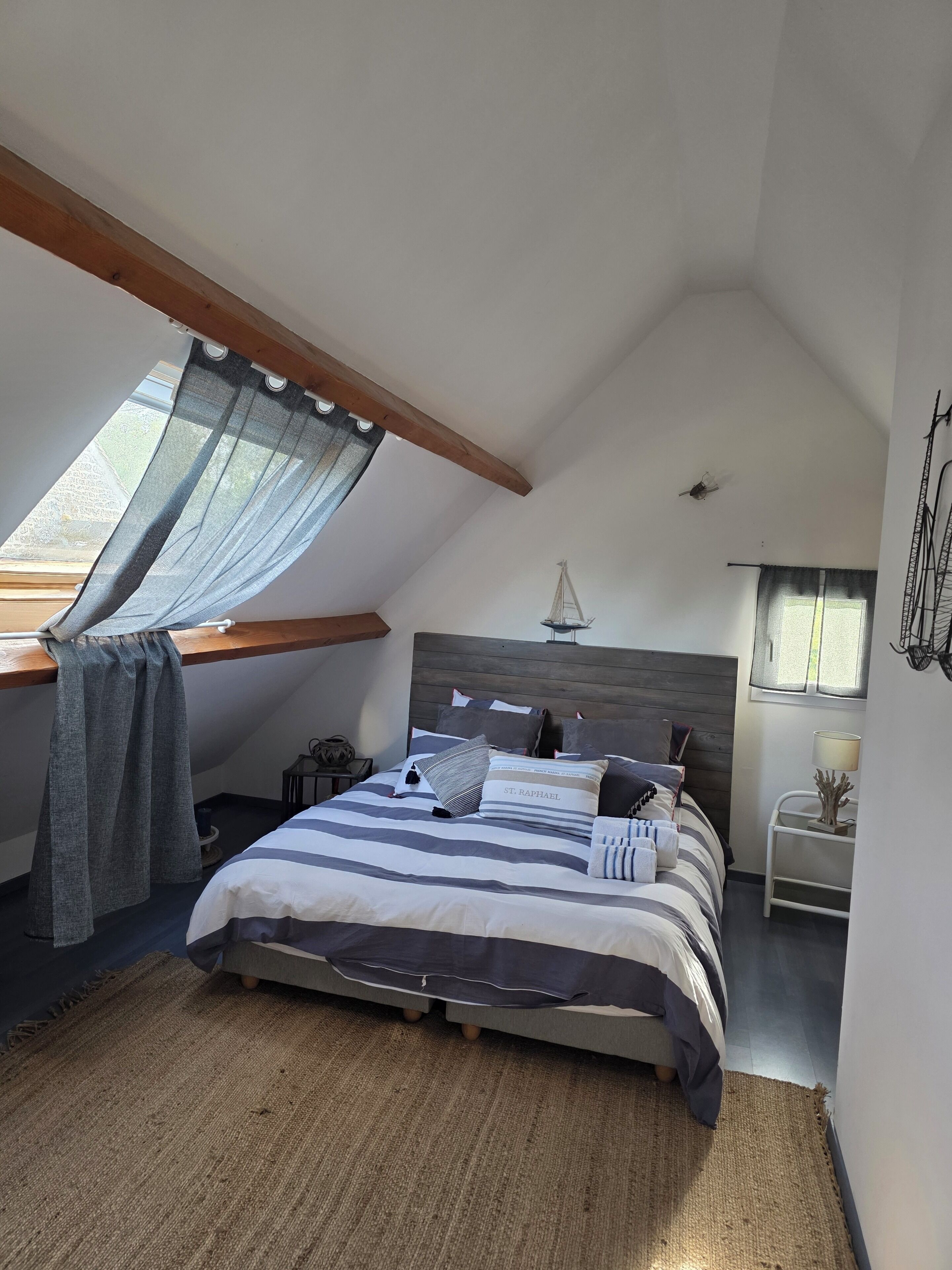 Quarto com teto inclinado, cama com roupa de cama listrada, decoração de um veleiro pendurado e uma mesinha com abajur.