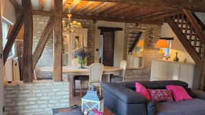 A rustic interior with exposed wooden beams, a stone wall, a dining area with a table and chairs, and a sofa with decorative pillows.