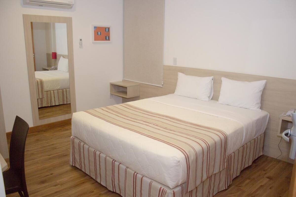 A hotel room with a bed, a desk, a chair, a nightstand, and a mirror.