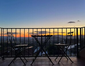 Ein Balkon mit zwei Stühlen und einem Tisch, mit Blick auf eine Skyline bei Sonnenuntergang.