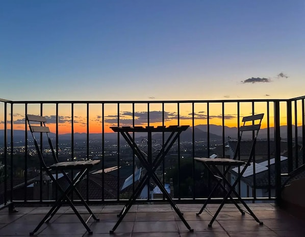 A balcony with two chairs and a table, overlooking a cityscape during sunset.