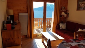 Quarto acolhedor com decoração em madeira, um sofá vermelho, uma mesa de madeira e vista para montanhas através de uma porta de vidro.