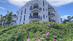 Un edificio blanco de varios pisos con balcones, rodeado de exuberante vegetación y flores rosadas.