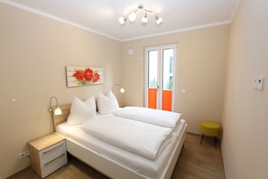 Ein Schlafzimmer mit Bett, Nachttischen, einer Lampe, einem kleinen Hocker und einem Fenster mit Jalousien.
