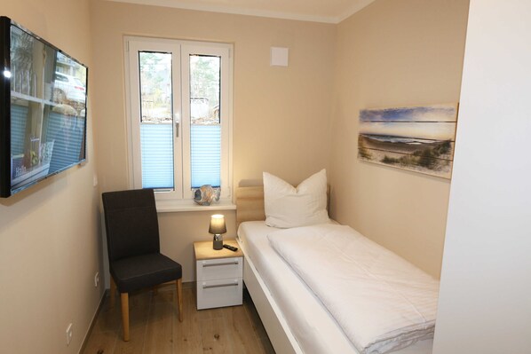 Une petite chambre à coucher, bien rangée, avec un lit simple, une chaise, une table de chevet avec une lampe et une fenêtre équipée de stores.