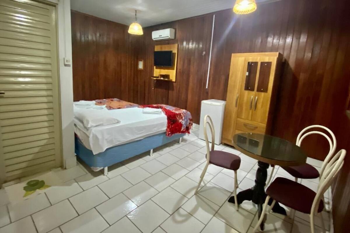 Quarto com cama, uma mesa com cadeiras, guarda-roupa de madeira e televisão na parede.