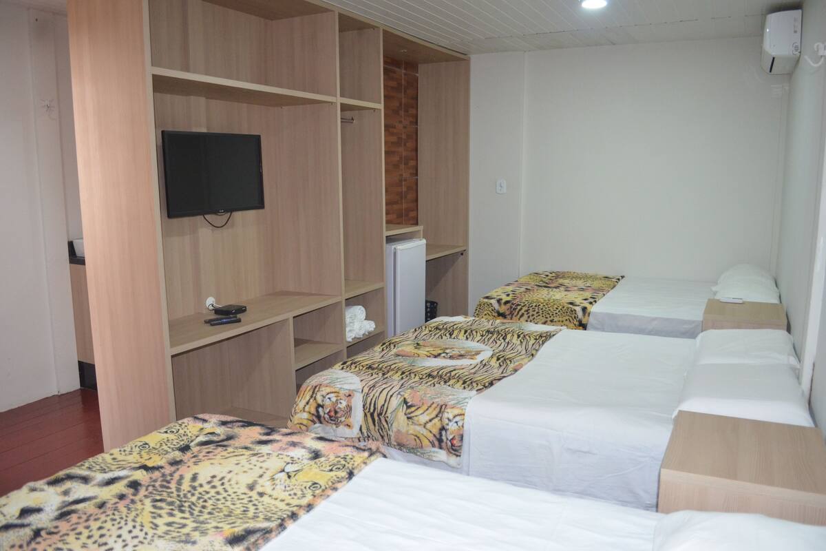Quarto de hotel com três camas de solteiro, uma TV fixada em um guarda-roupa de madeira e uma pequena geladeira branca.
