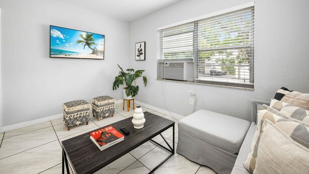 A modern living room with a flat-screen TV, a large window with blinds, a sofa, a coffee table with a magazine, and two patterned ottomans. - HAVANA5 | spacious escape in miami's cultural hub (Miami)