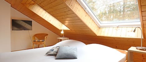 Une chambre avec un plafond en pente, un lit, une chaise, un téléviseur et une fenêtre donnant sur des arbres.
