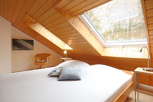Un dormitorio con techo inclinado, una cama, una silla, un televisor y una ventana con vistas a árboles.