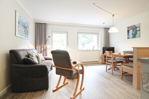 A living room with a sofa, a rocking chair, a dining table with chairs, a TV, and a window with curtains.