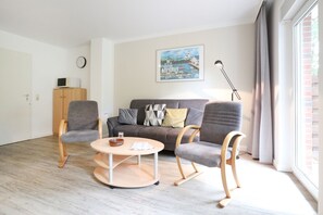 A modern living room with a grey sofa, two wooden chairs, a round coffee table, and a painting of a coastal scene. - Vacationer village 27c, Germany (Ostseebad Boltenhagen)