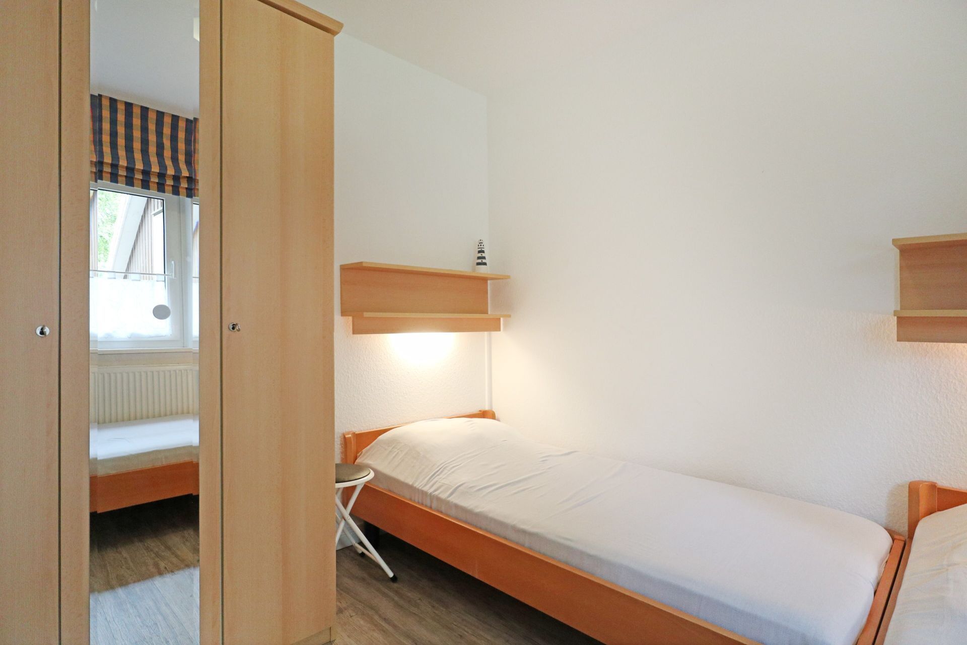 A small, single-bed room with a wooden bed frame, a small bedside table, and a window with a striped blind.