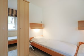 A small, single-bed room with a wooden bed frame, a small bedside table, and a window with a striped blind.