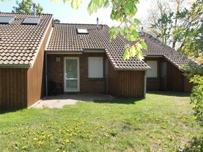A two-story house with a tiled roof, a brick entrance, and a small front yard with grass and dandelions. - Vacationer village 27c, Germany (Ostseebad Boltenhagen)