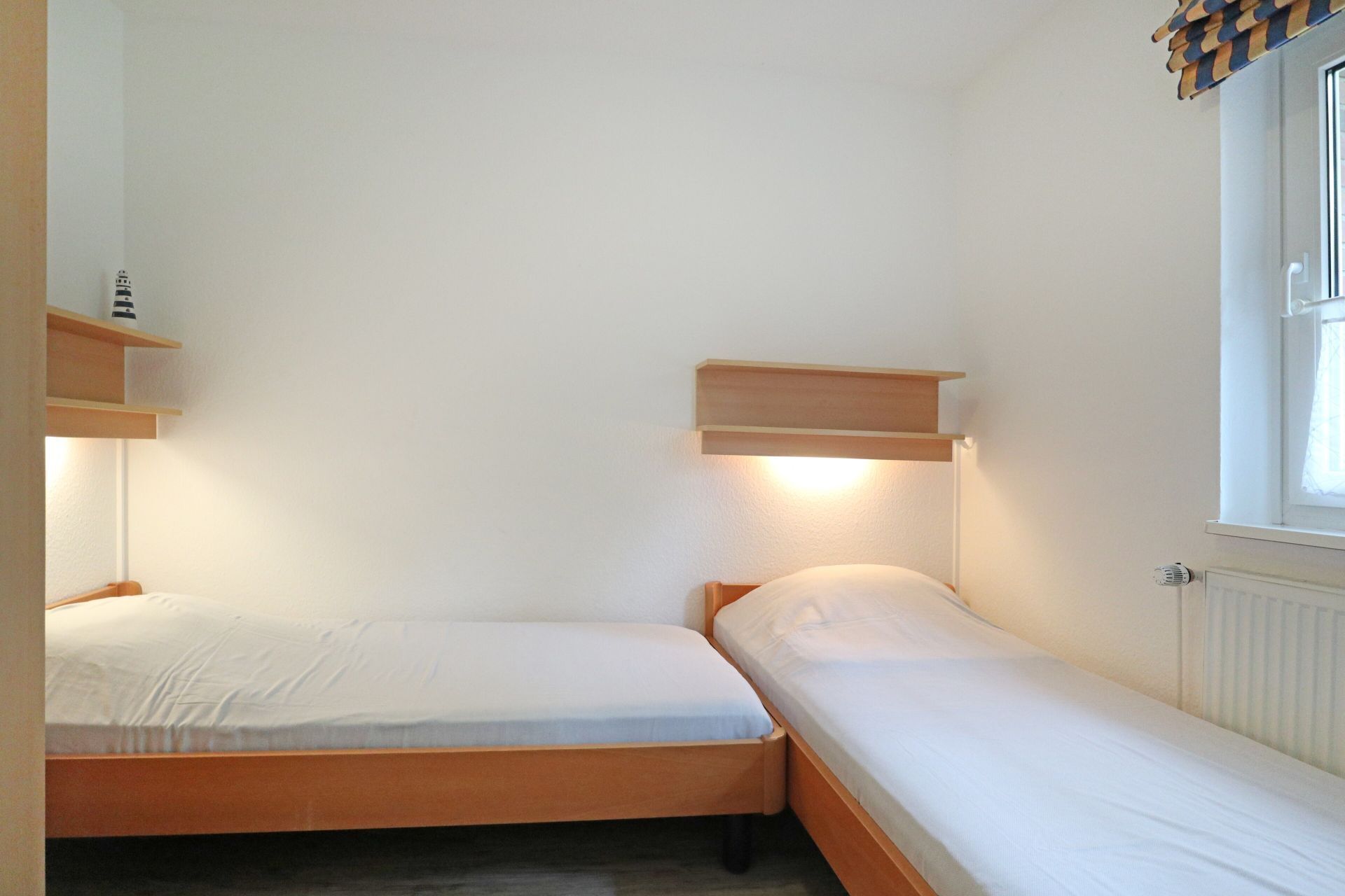 Un petit logement composé d’une seule pièce, comprenant deux lits simples, une fenêtre avec stores et un radiateur.