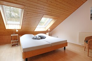 A bedroom with a wooden sloped ceiling, two skylights, a bed with pillows, bedside tables, and a wicker chair. - Vacationer village 27c, Germany (Ostseebad Boltenhagen)