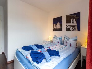 Ein ordentlich bezogenes Bett mit blau-weißer Bettwäsche, zwei Nachttische mit Lampen und zwei an der Wand hängende Bilder von Gebäuden.