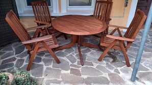 Une table ronde en bois, entourée de quatre chaises, est placée sur une terrasse en pierre. On aperçoit une porte et des fenêtres à l’arrière-plan.