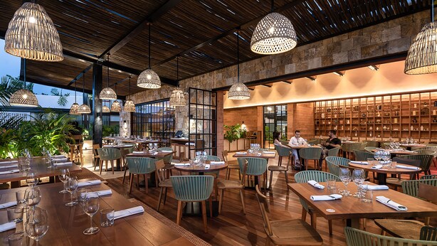 A restaurant with wooden tables and chairs, hanging pendant lights, and a brick wall with shelves. - Las Brisas Merida (Mérida)