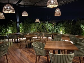 Un área de comedor al aire libre con mesas y sillas de madera, rodeada de exuberante follaje e iluminada por lámparas colgantes.