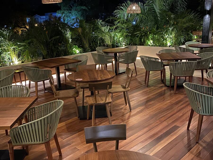 An outdoor dining area with wooden tables and chairs, surrounded by lush greenery and illuminated by hanging lamps.