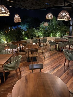 Un espacio al aire libre para comer con mesas y sillas de madera, rodeado de abundante follaje e iluminado por lámparas colgantes.