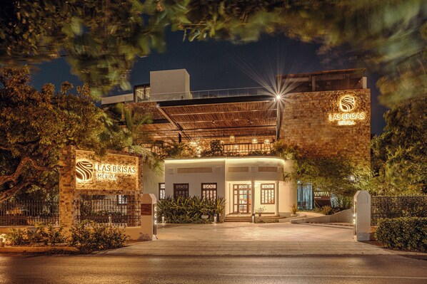 Entrada de un hotel con un letrero que dice "Las Brisas Merida", rodeada de follaje y un camino bien iluminado.