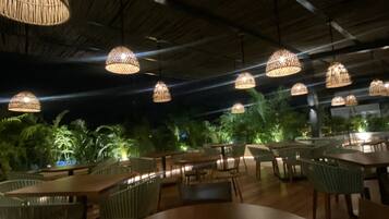 Un comedor interior con mesas y sillas redondas, lámparas de mimbre colgantes y vistas a palmeras afuera.