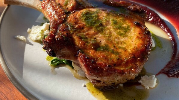 A close-up of a cooked pork chop with a glossy, herb-topped crust, served on a white plate with a creamy sauce and some greens. - Las Brisas Merida (Mérida)