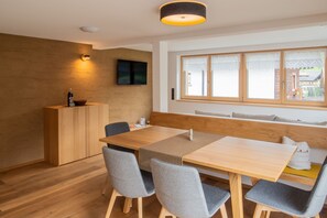 Un comedor moderno con una mesa de madera, sillas grises, un banco de madera, un televisor montado en la pared y una ventana con cortinas.