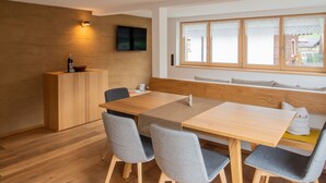 A modern dining area with a wooden table, grey chairs, a wooden bench, a television mounted on the wall, and a window with curtains. - Vacation apartment Gutgehen (1-2 persons) - Moosbrugger Rosmarie - Haus WohlSEIN (Schoppernau)