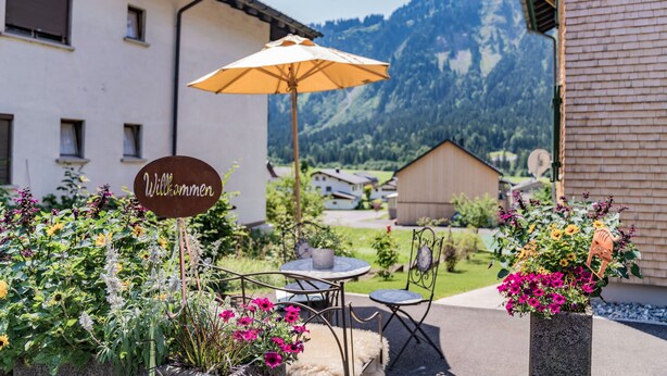 A patio with outdoor seating, a welcome sign, and a mountain backdrop. - Vacation apartment Gutgehen (1-2 persons) - Moosbrugger Rosmarie - Haus WohlSEIN (Schoppernau)
