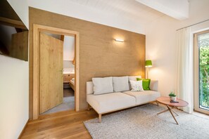 A modern living room with a wooden accent wall, a sofa, a small table with a plant, and a view through a window. - Vacation apartment Enjoy (2-4 persons) - Moosbrugger Rosmarie - Haus WohlSEIN (Schoppernau)
