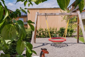 A wooden swing set with a red tire swing, surrounded by greenery and a building with a wooden fence in the background.