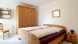 Un dormitorio con una cama de madera, un armario de madera, una silla, una mesita de noche y un pequeño estante con libros y una planta.