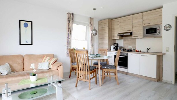 Una sala moderna con un sofá, una mesa de centro de vidrio y un comedor con sillas de madera. Se ve una cocina con armarios blancos y un microondas al fondo.